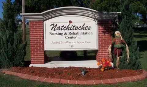 Brick entrance sign reads Natchitoches Nursing and Rehabilitation Center with landscaping and a scarecrow.