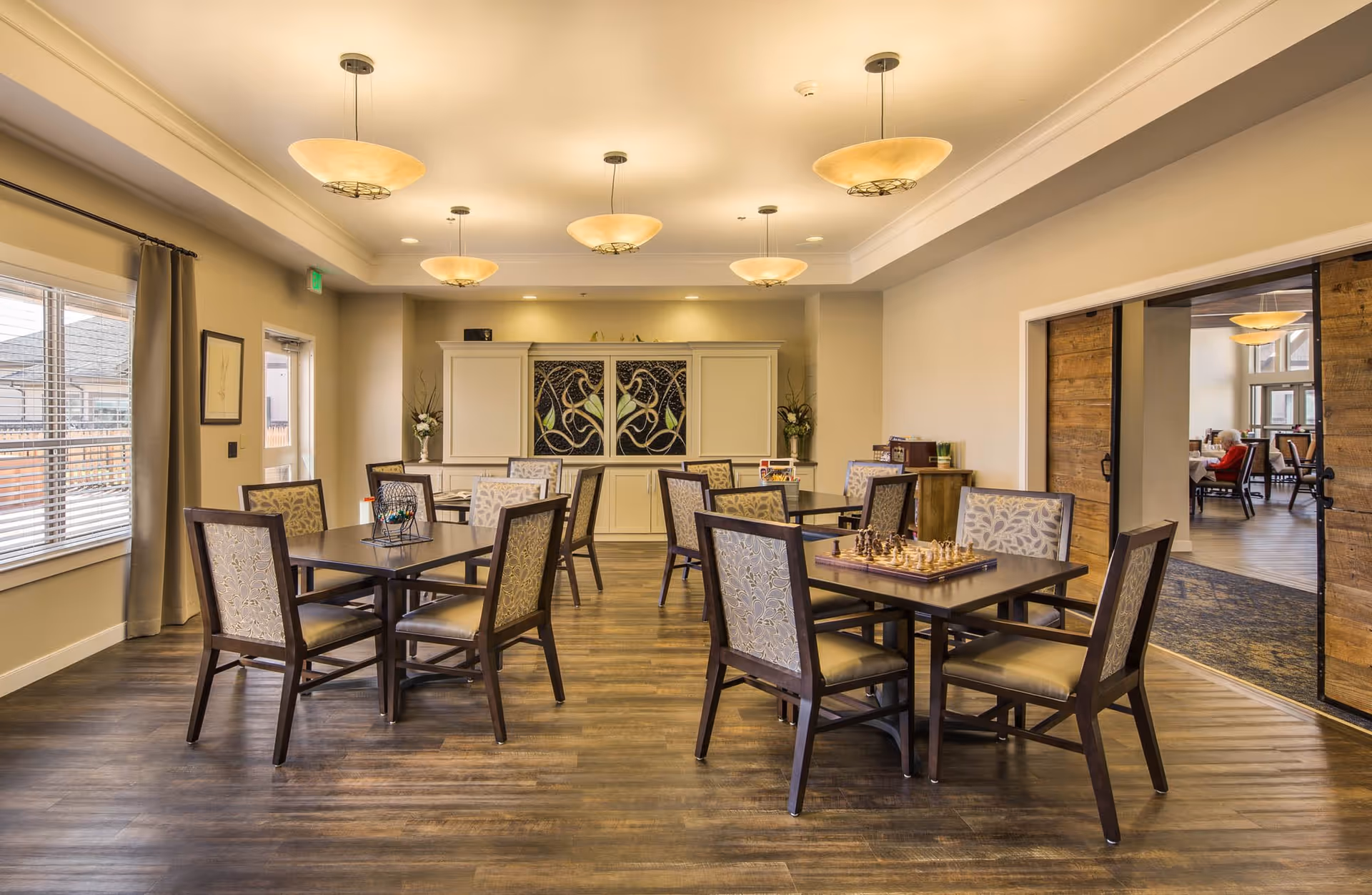 A well-lit dining room with several square tables and cushioned chairs arranged neatly on a wooden floor. The room features warm beige walls, ceiling lights, and a large window with curtains on the left side. A decorative cabinet with artistic panels is positioned against the far wall. Through an open doorway with sliding wooden barn doors, another room with more tables and chairs is visible, where an elderly person is seated.