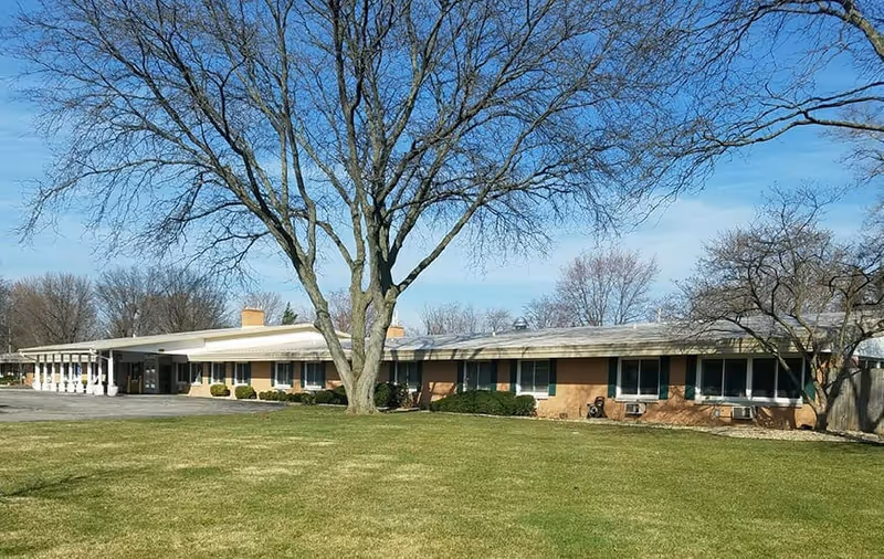 Single-story brick building with multiple windows and green shutters, surrounded by a large grassy lawn and leafless trees under a clear blue sky.