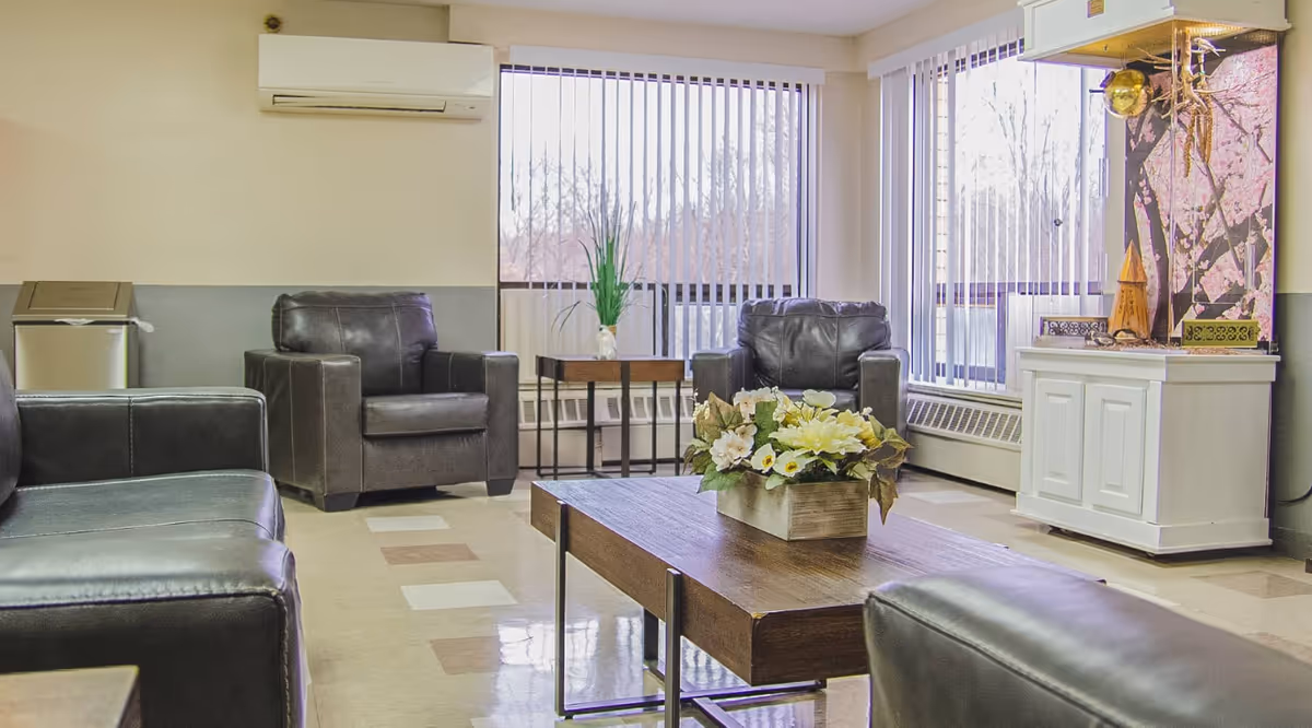 A bright living room area with large windows covered by vertical blinds. The room features black leather chairs and sofas arranged around a wooden coffee table with a floral centerpiece. There is a white cabinet with decorative items and an air conditioning unit mounted on the wall.