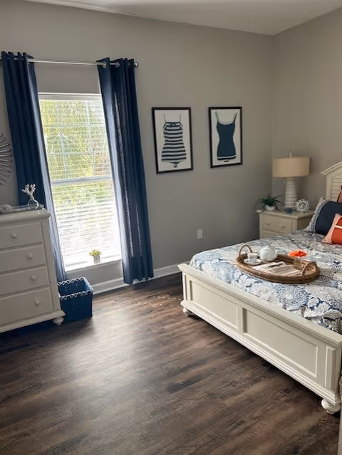 Sunlit bedroom with a white wooden bed and dresser, dark curtains at a window, and framed swimsuit artwork on a gray wall.