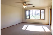 Empty living room with a ceiling fan, large picture window letting in sunlight, and a carpeted floor.