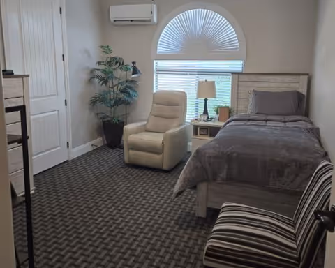A bedroom with a single bed covered in gray bedding, a beige recliner chair, a small nightstand with a lamp and a plant, a large window with blinds and a decorative half-circle window above it, a potted plant in the corner, and a striped chair near the door.
