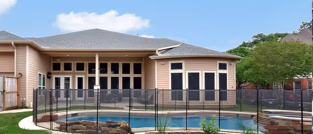 View of the back exterior of a single-story residential building with large windows and a covered patio area. In the foreground, there is a fenced swimming pool surrounded by a stone border and some greenery. Trees and neighboring houses are visible in the background under a partly cloudy sky.