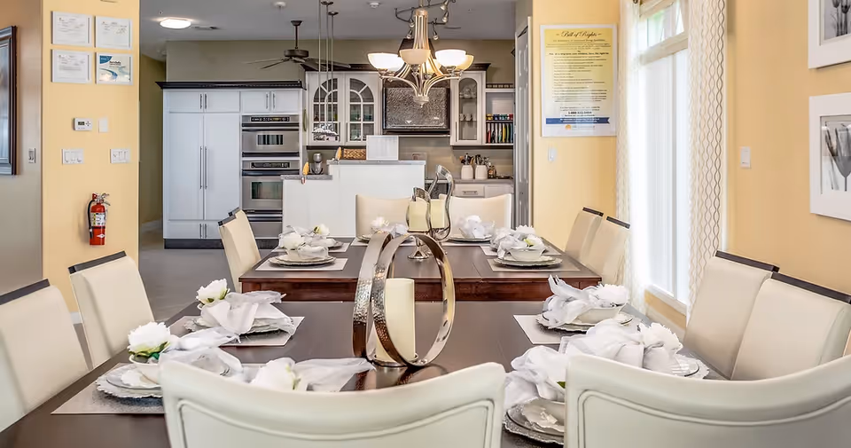 A formal dining room with a long set table and upholstered chairs, place settings, decorative centerpiece, and a view into a modern kitchen.