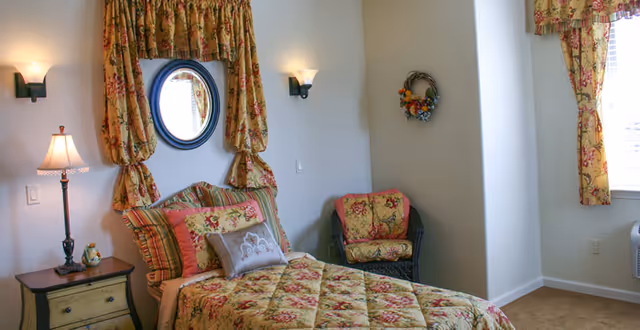 A cozy bedroom with a single bed covered in a floral patterned quilt and matching pillows. The bed is positioned against a wall with a round mirror above it, flanked by two wall sconces. To the left of the bed is a small wooden nightstand with a lamp and a decorative item. In the corner, there is a cushioned chair with a floral cushion matching the bedspread. The room has light-colored walls and a window with floral curtains allowing natural light to enter.
