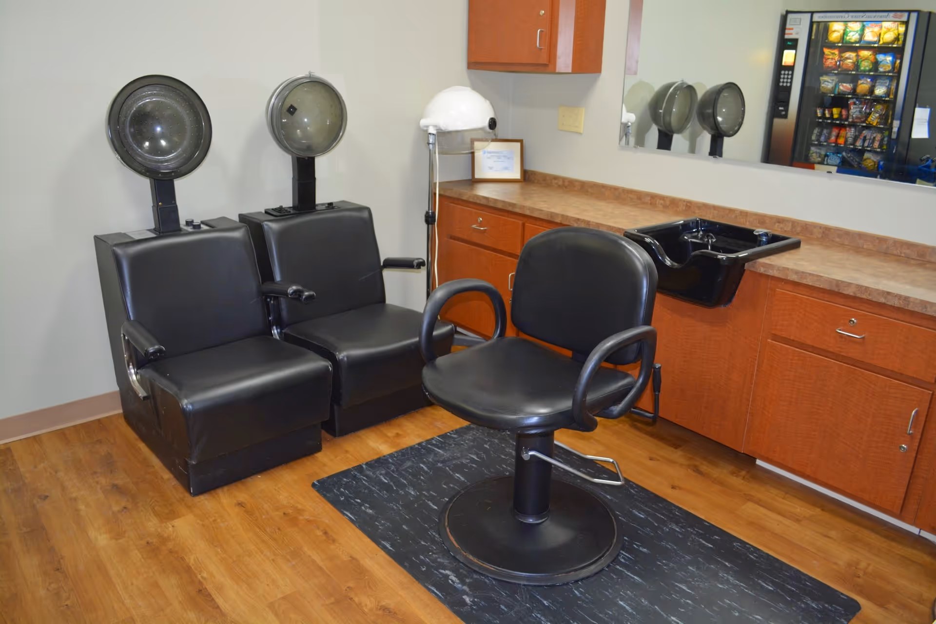 Interior of a hair salon area with two black leather hair drying chairs, a black salon styling chair on a black mat, a black shampoo sink, wooden cabinets with a countertop, a large mirror, and a vending machine reflected in the mirror.