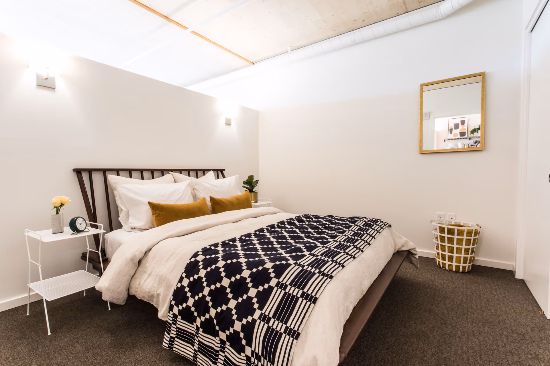 A modern bedroom with a wooden bed frame, white bedding, two mustard yellow pillows, and a black and white patterned throw blanket. There are two white side tables, one with a small plant and the other with a clock and a flower vase. A square mirror hangs on the wall above a laundry basket, and the room has white walls and carpeted flooring.