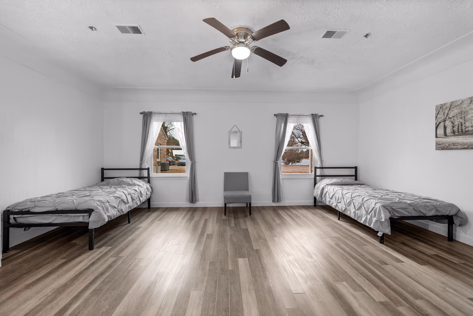 A simple bedroom with two single beds placed against opposite walls, each with gray bedding. Between the beds is a gray chair centered under a small wall mirror. Two windows with sheer white curtains and gray drapes allow natural light into the room. The floor is wood laminate, and a ceiling fan with a light is mounted on the white ceiling. A black and white framed picture hangs on the right wall.