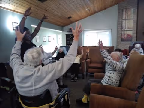A group of elderly individuals seated in a common room with wooden ceiling and light green walls, participating in a group activity with their arms raised. The room has comfortable chairs and a window with blinds letting in natural light.
