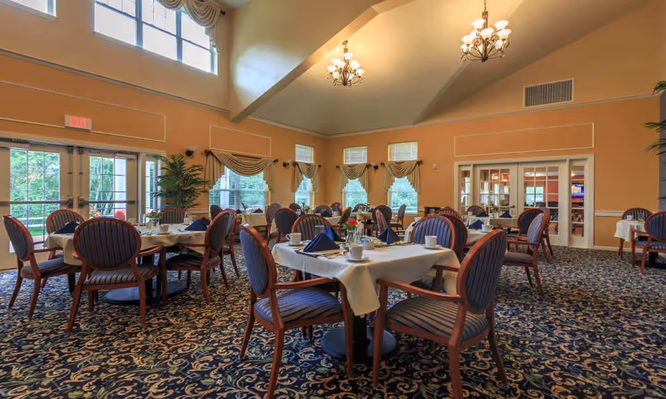 A spacious dining room with multiple round tables covered with white tablecloths, each set with blue napkins, cups, and small flower arrangements. The room features large windows with decorative curtains, high ceilings with chandeliers, and patterned carpet flooring. There are wooden chairs with blue and red striped cushions around the tables.