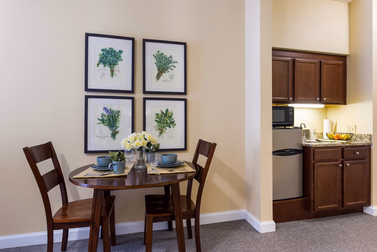 A small dining area with a round wooden table set for two with blue dishes, glasses, and a vase of white flowers. On the wall above the table are four framed botanical prints. To the right is a kitchenette with dark wooden cabinets, a small refrigerator, a microwave, a sink, and a granite countertop with a bowl of fruit.