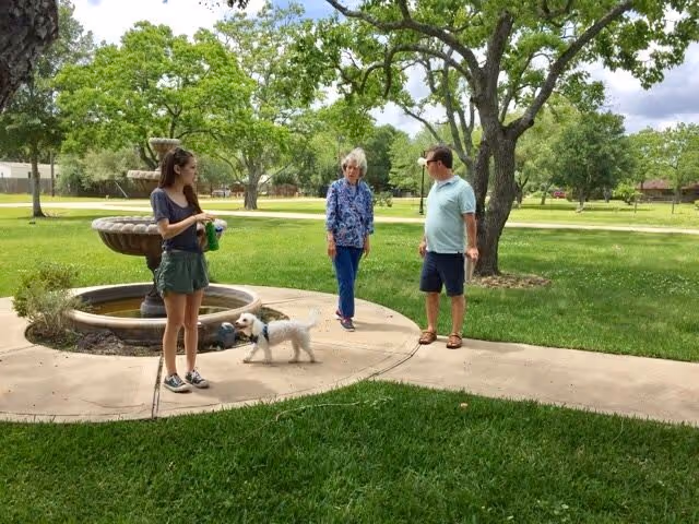 Three people standing on a paved walkway in a green park-like outdoor area with trees and a water fountain. One person is holding a small white dog on a leash.