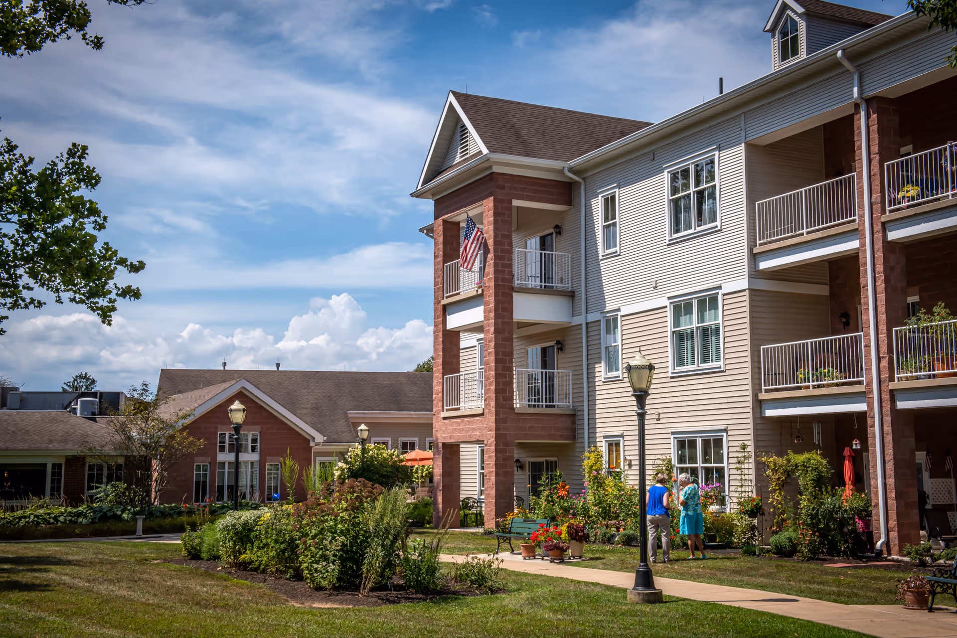 Exterior view of a senior living community building with balconies, landscaped gardens, and a walkway where two people are standing and talking. The sky is partly cloudy and the area is well-maintained with greenery and flowers.