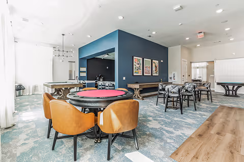 Open, bright communal activity and dining room with multiple tables and chairs, including a round poker table with red felt and a blue accent wall.