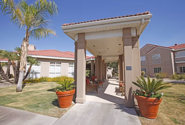 Outdoor covered walkway with seating area at a senior living facility. The walkway is supported by beige columns and has a tiled roof. There are large potted plants on either side of the walkway entrance, palm trees, and well-maintained grass and shrubs surrounding the area under a clear blue sky.