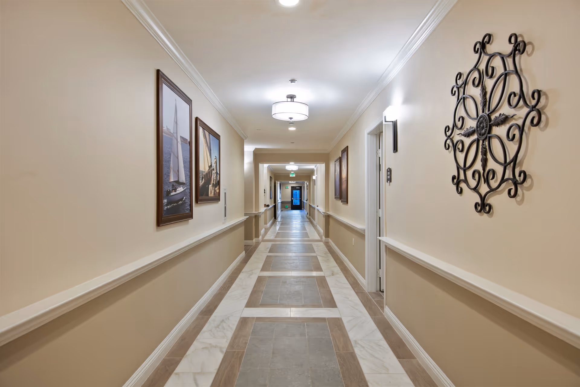 A long, well-lit hallway in a senior living facility with beige walls, decorative wall art, framed pictures of sailboats, and a patterned tile floor. The hallway has ceiling lights and wall sconces, with doors along the right side and an exit door visible at the far end.