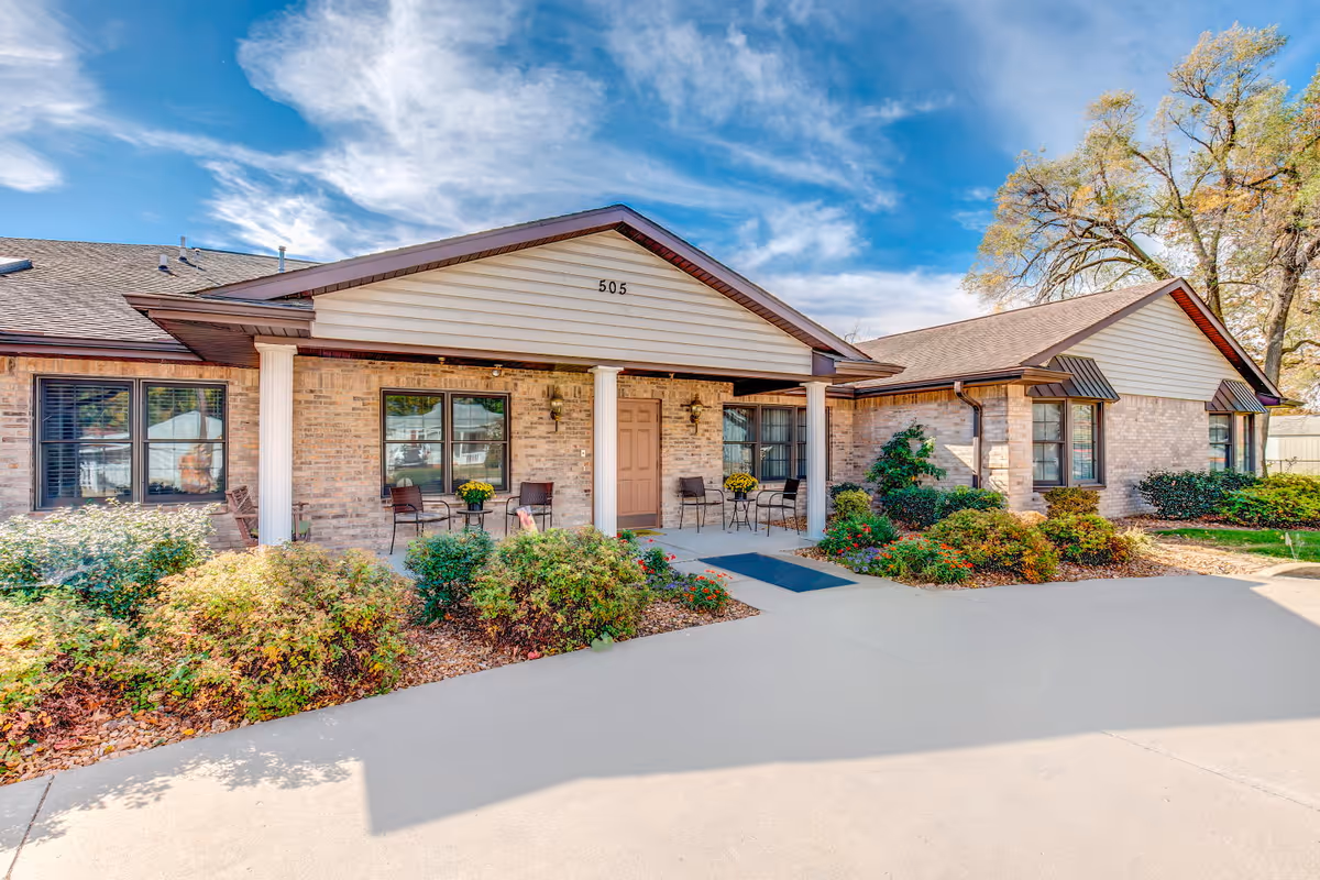 Exterior view of a single-story brick building with a covered entrance supported by white columns. The building has several windows with dark frames and a door under the covered porch. There are chairs and small tables with potted plants on the porch. Shrubs and greenery surround the building, and the sky is partly cloudy with blue patches.