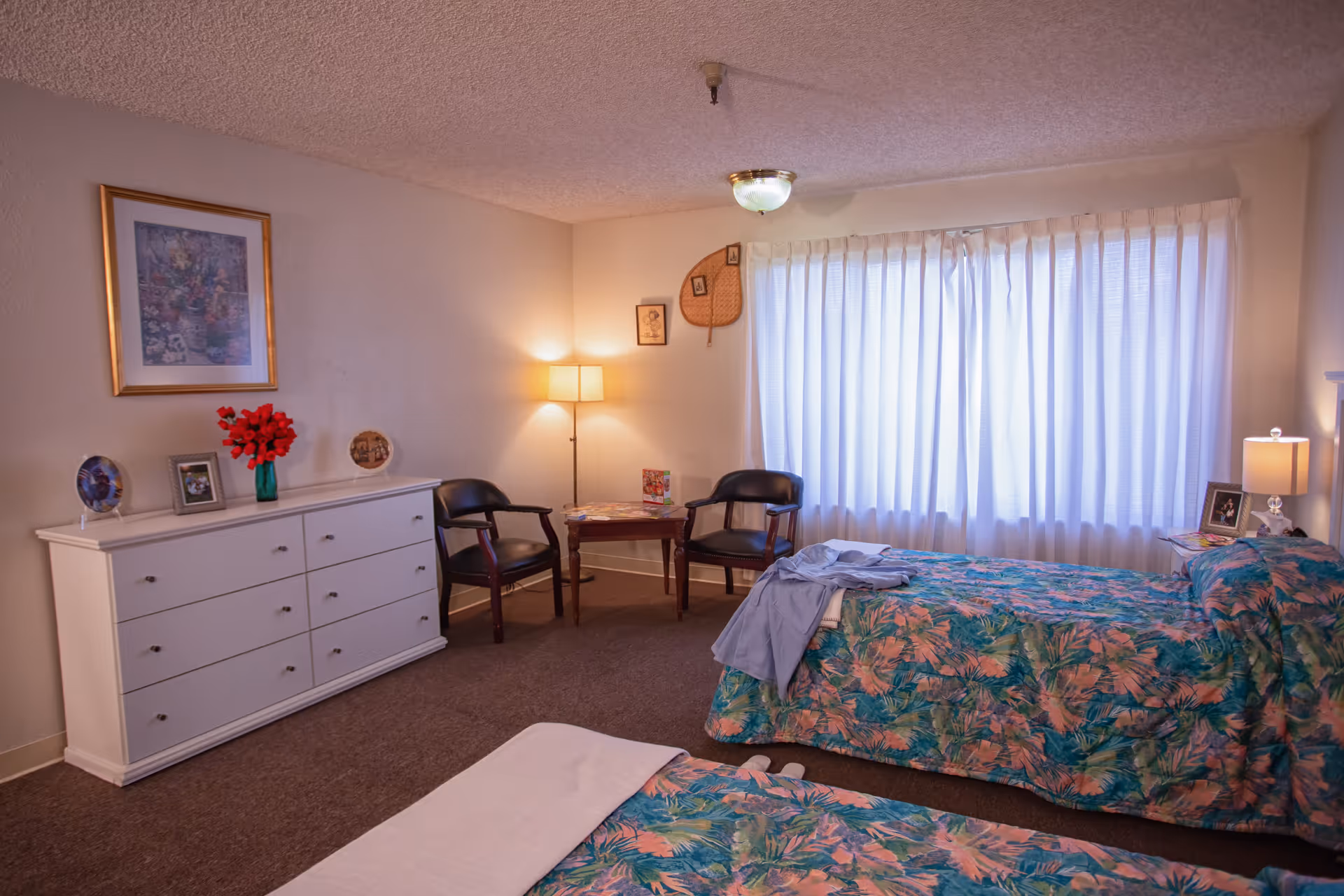 A cozy bedroom with two beds covered in floral patterned bedspreads. There is a white dresser with decorative items and framed pictures on top. Two black chairs and a small wooden table are positioned near a window with white curtains. The room is softly lit by a floor lamp and a ceiling light.
