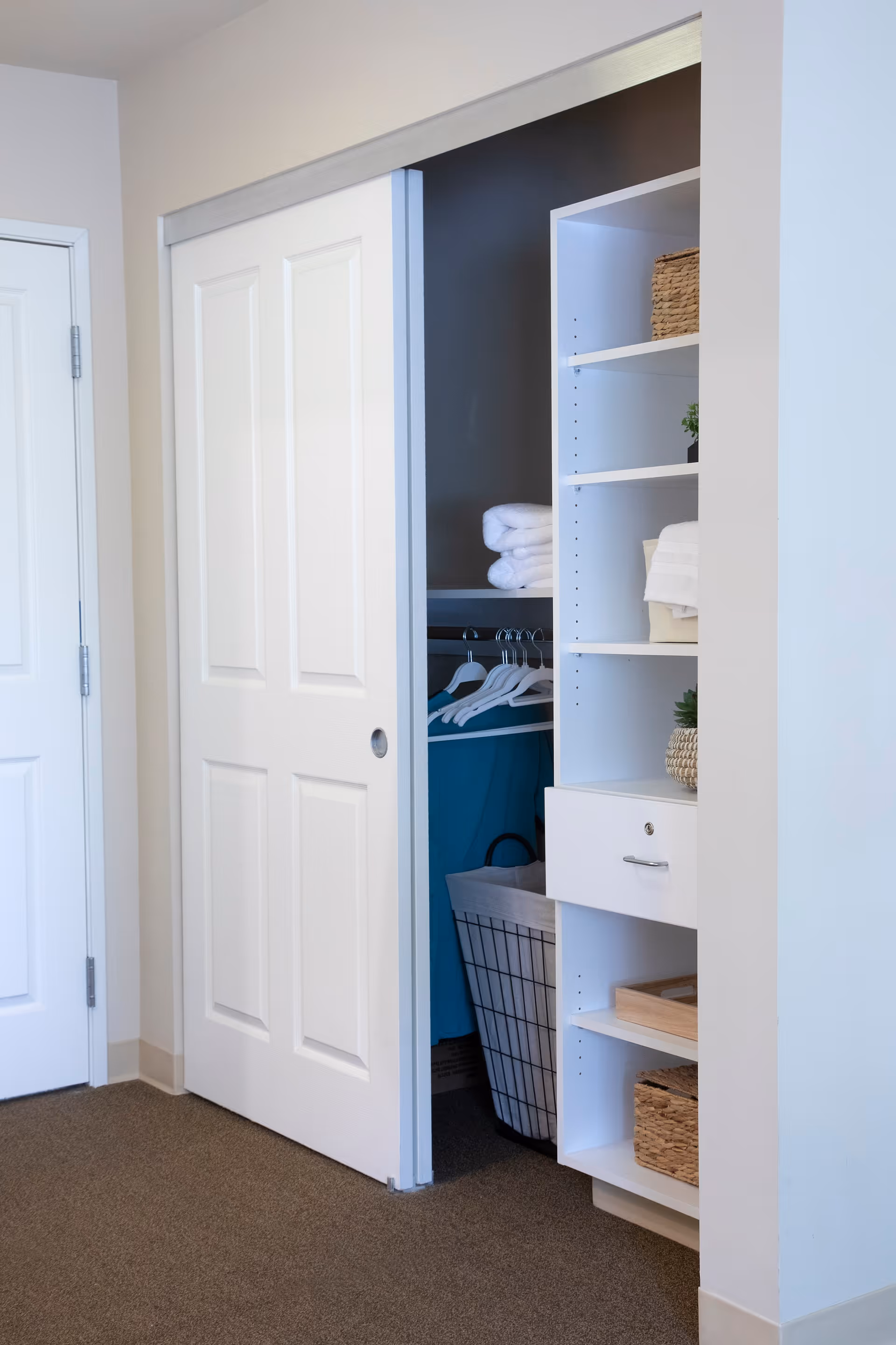 Interior view of a closet with a sliding white door partially open, revealing hanging clothes, folded towels, and a laundry basket inside. Next to the closet is a white shelving unit with baskets, folded towels, and small decorative plants.