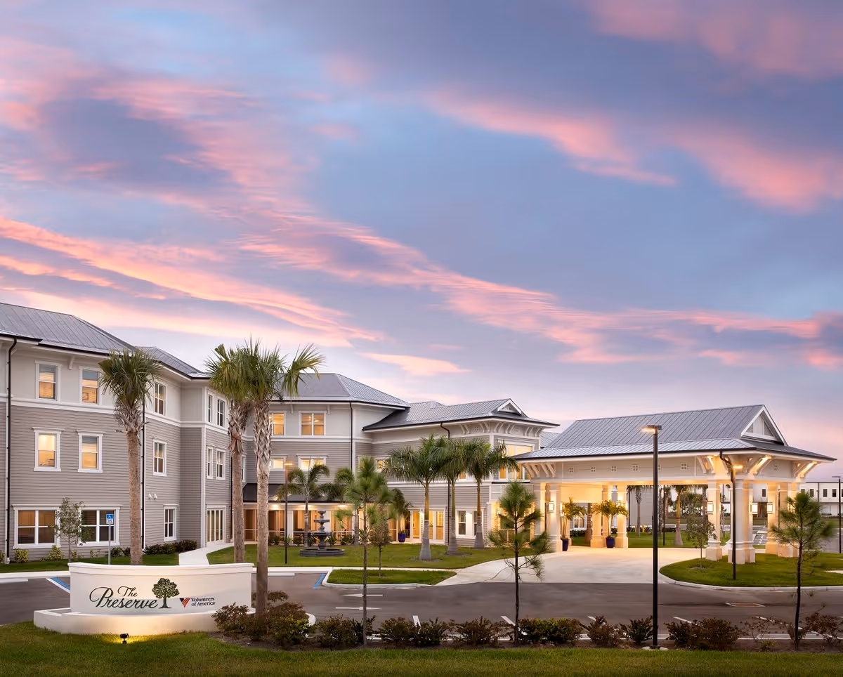 Exterior view of The Preserve in Fort Myers senior living facility at sunset, showing a large three-story building with palm trees, a covered entrance, and a landscaped driveway with a sign in front.