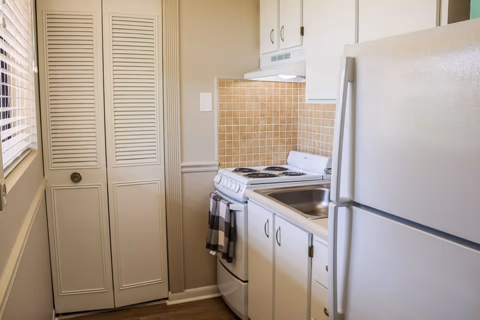 Small kitchen area with white cabinets, a white refrigerator, a white stove with four burners, a stainless steel sink, and beige tiled backsplash. There is a window with blinds on the left and a closed louvered door next to the stove.