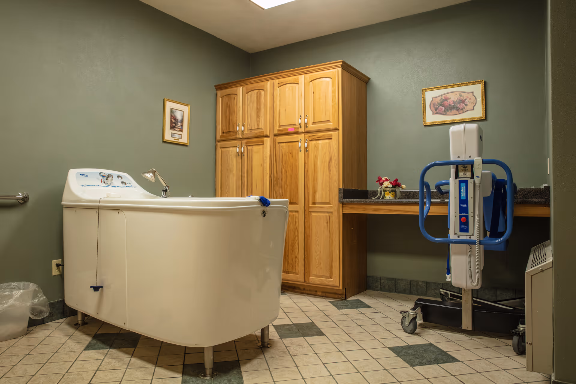 A bathroom with a large white bathtub, wooden cabinets, a countertop with a small flower arrangement, and a blue and white patient lift device. The walls are painted green and there are two framed pictures hanging on the walls.