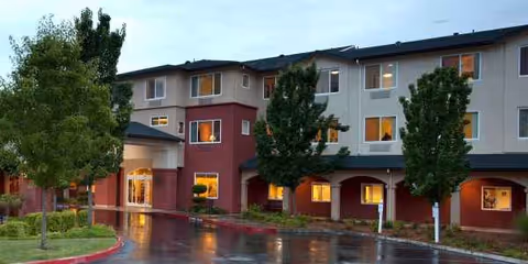 Two-story senior living building with red and beige walls, trees, and a wet parking lot.