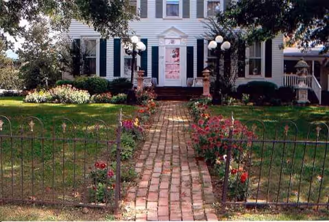 Brick walkway leads to a white house with pink front door, surrounded by flowers.