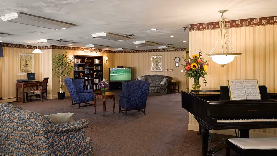 A senior living community lounge area with patterned sofas and armchairs arranged around a small wooden table with a flower vase. A black grand piano with sheet music and a flower arrangement on top is visible on the right. In the background, there is a bookshelf, a TV, a computer desk with a chair, and framed artwork on the walls. The room has striped wallpaper and carpeted floors.