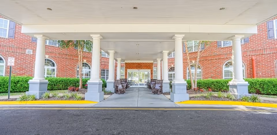Covered entrance area of a senior living facility with white columns and brick walls. There are rocking chairs arranged along the sides under the covered walkway, with greenery and small trees planted along the building. The entrance doors are glass and located at the far end of the walkway.