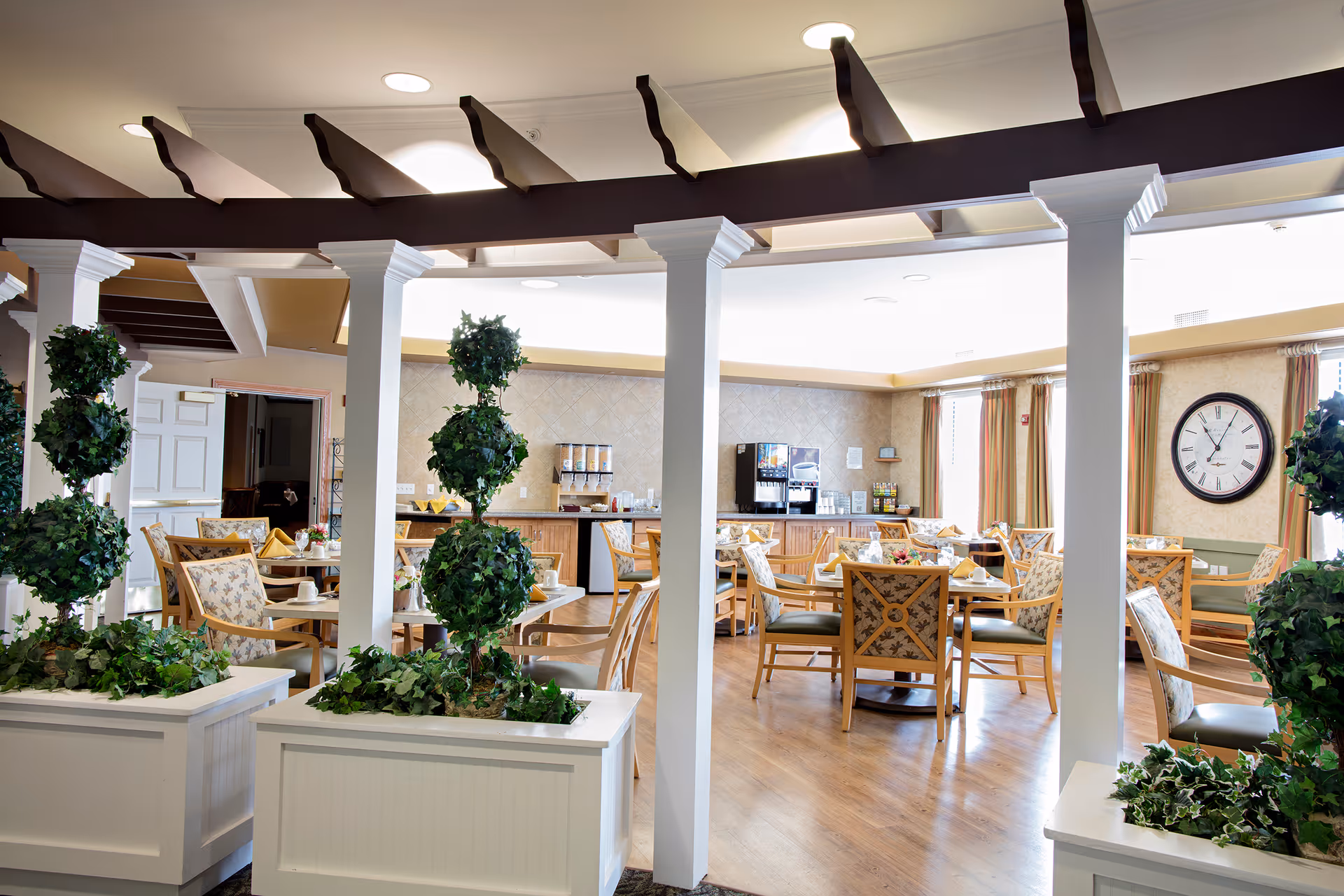A bright and inviting dining room in a senior living facility with wooden tables and chairs arranged neatly. The room features large windows with curtains, a wall clock, and a beverage station with coffee and juice dispensers. Decorative green plants in white planters and white columns add to the welcoming atmosphere.