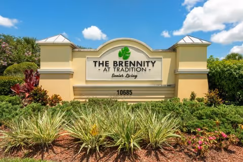 Entrance sign for The Brennity at Tradition Senior Living surrounded by landscaped greenery and plants under a blue sky with some clouds.