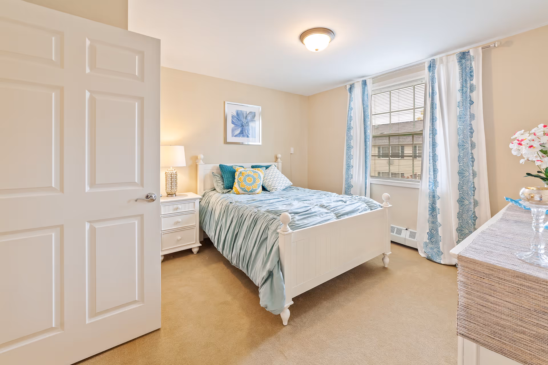 A bright and cozy bedroom with a white wooden bed frame, light blue bedding, and decorative pillows. There is a white nightstand with a lamp on the left side of the bed, a framed floral artwork on the wall above the bed, and a window with blue and white patterned curtains letting in natural light. A dresser with a vase of flowers is visible on the right side of the room.
