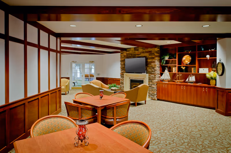 A cozy senior living common area featuring wooden tables with cushioned chairs, a stone fireplace with a mounted flat-screen TV above it, and built-in wooden shelves decorated with model sailboats and plants. The room has warm lighting and carpeted floors, with a seating area of armchairs near the fireplace and a door leading to another room in the background.