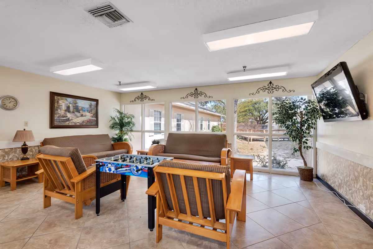 A bright and cozy common area in a senior living facility featuring two cushioned wooden sofas, two cushioned wooden armchairs, a foosball table in the center, a wall-mounted flat-screen TV, a large window with a view of the outdoors, a potted plant, a framed painting, a wall clock, and a table lamp on a side table.