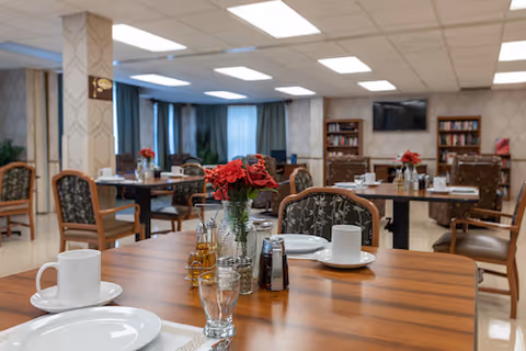 A dining room with wooden tables and chairs arranged neatly. Each table has white plates, cups, glasses, and condiments. A vase with red flowers is placed in the center of the closest table. The room has patterned wallpaper, bookshelves, and a television mounted on the wall.
