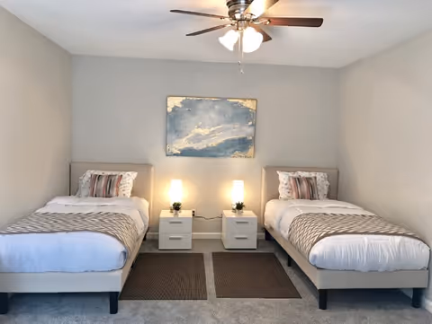 A bedroom with two single beds separated by two white nightstands, each with a small lamp and a small plant. Each bed has white bedding, a patterned throw blanket, and a decorative pillow. Above the nightstands is a large abstract painting in blue and gold tones. The room has a ceiling fan with lights and neutral-colored walls and carpet.