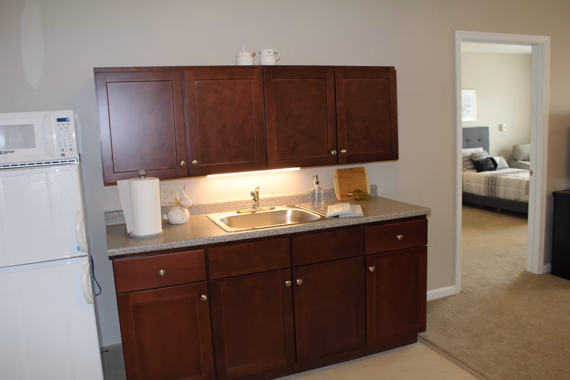 A small kitchen area with dark wooden cabinets, a countertop with a sink, a paper towel holder, and a soap dispenser. A white microwave sits on top of a white refrigerator to the left. Through an open doorway on the right, a bedroom with a bed and pillows is visible.