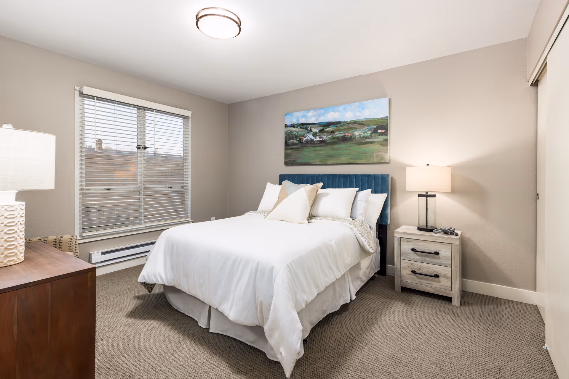 A neatly made bed with white bedding and multiple pillows in a bedroom. The bed has a blue upholstered headboard. There is a wooden nightstand with a lamp and some decorative items on the right side of the bed. A large window with blinds is on the left wall, and a landscape painting hangs above the bed. The room has beige walls and carpeted floor.
