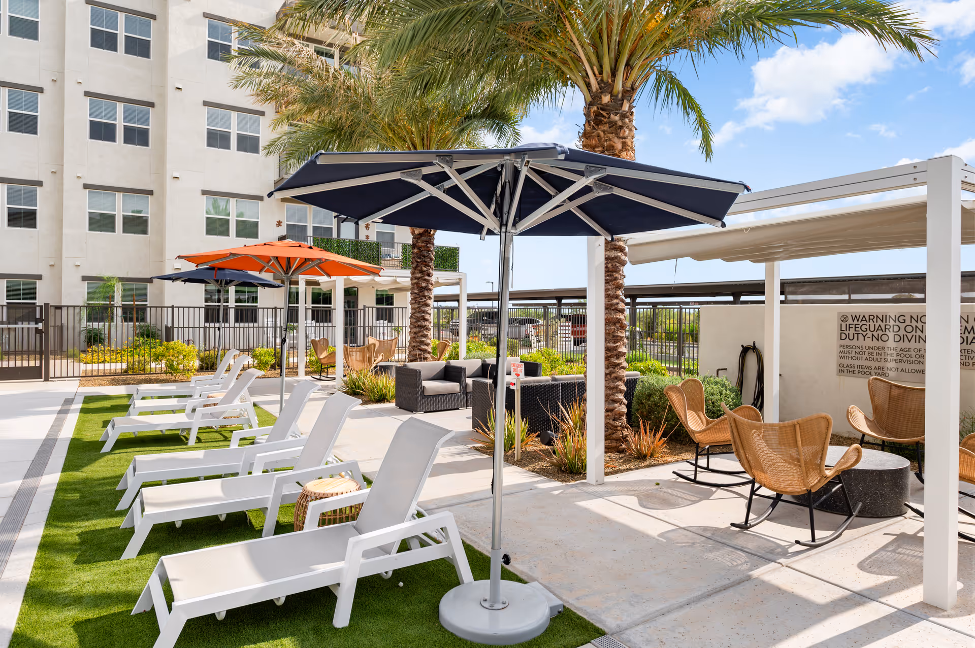 Outdoor seating area at Sage Mesa facility featuring white lounge chairs with umbrellas on artificial grass, palm trees, wicker chairs around a small table under a pergola, and a multi-story building in the background under a partly cloudy sky.