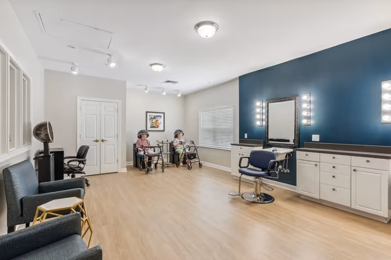 A spacious salon room in a senior living facility with light wood flooring and white walls, one wall painted dark blue. The room contains salon chairs, hair dryers, and two elderly women sitting under hair dryers with walkers beside them. There are cabinets with mirrors and lights on the blue wall, and comfortable seating with small tables on the left side.