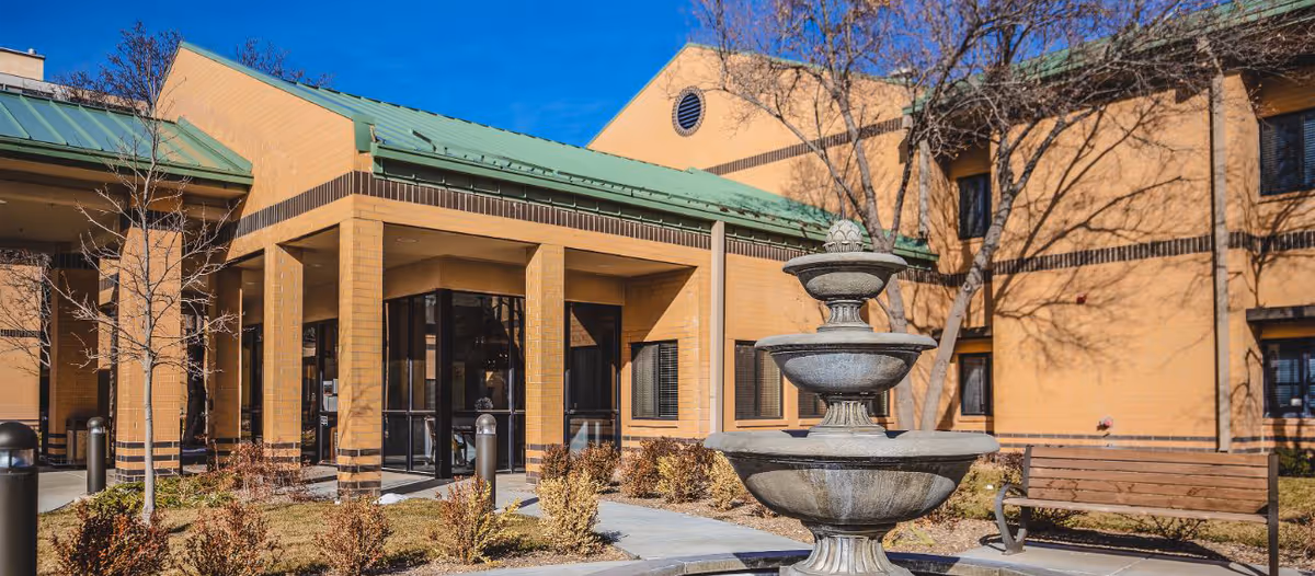 Exterior view of a senior living facility building with a green metal roof, beige brick walls, and large windows. In the foreground, there is a three-tiered stone fountain surrounded by a circular stone border, with a wooden bench and leafless trees nearby under a clear blue sky.