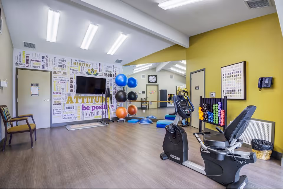 A bright exercise room in an assisted living facility featuring a recumbent exercise bike, colorful dumbbells on a rack, large exercise balls, balance pads, and a wall with motivational words like 'ATTITUDE', 'be positive', and 'dream big'. The room has wood flooring, a large mirror, and fluorescent ceiling lights.