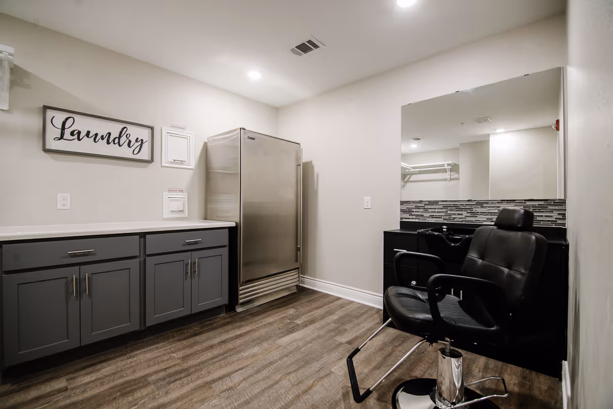 Interior room with gray cabinets, a stainless steel refrigerator, a black salon chair in front of a large mirror, and a sign on the wall that says 'Laundry'. The room has wood flooring and white walls with recessed lighting.