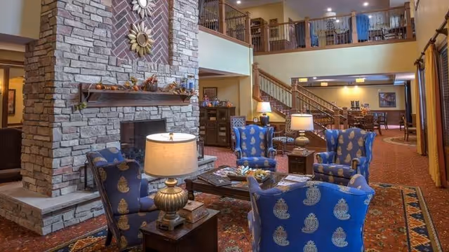 A cozy senior living facility common area with a large stone fireplace decorated with autumn-themed items. Four blue armchairs with gold patterns are arranged around a wooden coffee table with magazines. Two table lamps provide warm lighting. In the background, there is a staircase leading to an upper level with additional seating and a dining area.