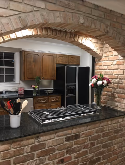 Kitchen area with a brick archway and brick walls, featuring a black countertop with a built-in gas stove. Wooden cabinets and a black refrigerator are visible in the background. Kitchen utensils are in a white container on the counter, and two vases with flowers are placed on the countertop and near the cabinets.