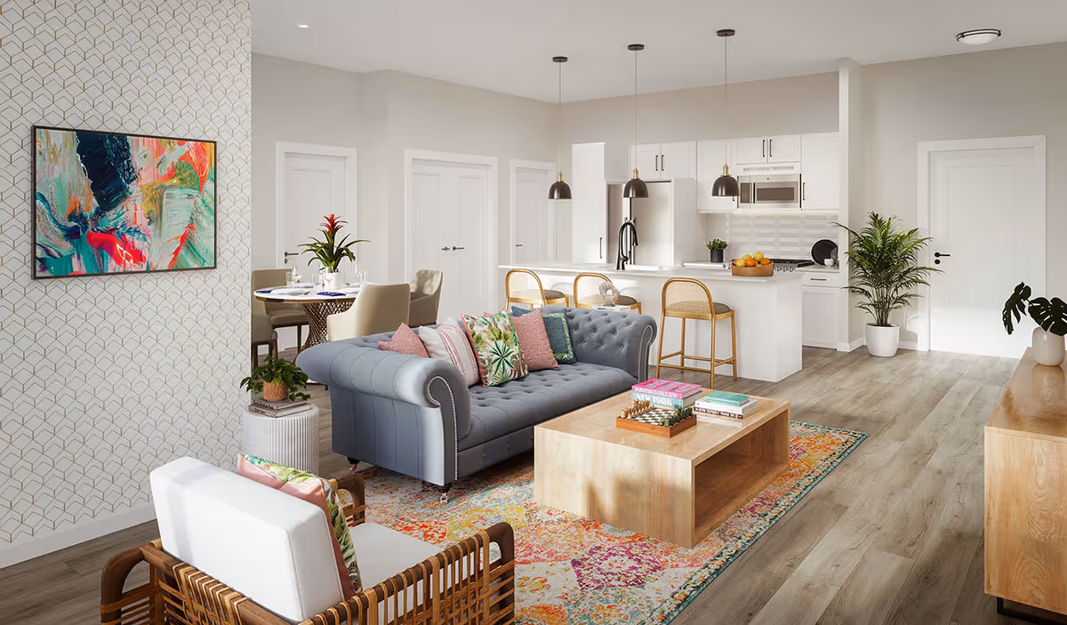 A bright and modern open-concept living room and kitchen area. The living room features a tufted gray sofa with colorful patterned pillows, a wooden coffee table with books and a chess set, and a wicker armchair with a white cushion and a decorative pillow. The kitchen has white cabinetry, a large island with three wooden bar stools, pendant lights, and stainless steel appliances. A round dining table with beige chairs and a plant centerpiece is visible in the background. The space is decorated with plants, a colorful abstract painting, and a patterned area rug on light wood flooring.