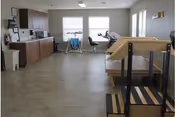 Bright multipurpose interior room with a kitchenette along the left wall, exercise equipment by the windows, and parallel walking bars in the foreground.