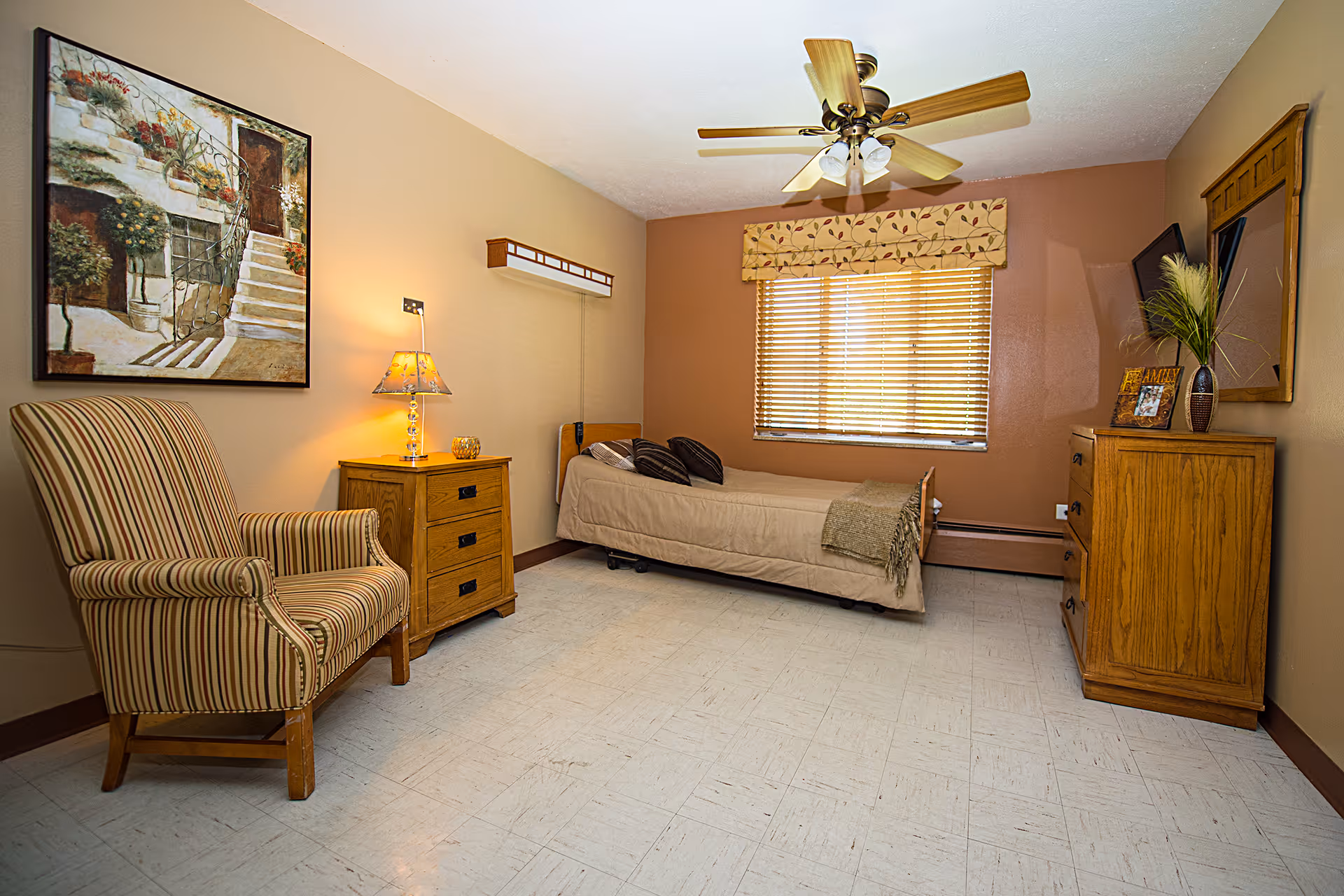 A simple bedroom with a single bed covered in beige bedding and two dark pillows. There is a wooden nightstand with a lamp and decorative item next to the bed. A striped armchair is positioned near the nightstand. On the opposite side of the room, there is a wooden dresser with a framed photo, a vase with plants, and a mirror above it. The window has wooden blinds and a floral valance. The ceiling has a fan with lights. The walls are painted in warm beige and brown tones, and the floor is light-colored tile.